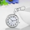 A close-up shot of a silver fob watch with a white face, attached to a silver carabiner clip. The watch is resting on a white surface, with a blurred white bowl-like object in the background and some blurred green and blue foliage. Produktbild-Vorschau