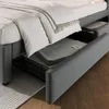 A close-up view of a grey upholstered bed frame with storage drawers pulled out, revealing folded blankets inside. Náhled obrázku produktu