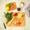 A top-down view of a collection of ingredients and kitchen tools arranged on a marble-like surface, including cutting boards, vegetables, herbs, and salmon. Anteprima dell’immagine prodotto
