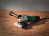 An angle grinder is placed on a wooden surface with a concrete wall in the background. Produktbild-Vorschau