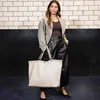 A woman stands in front of a brick wall and a black textured wall, holding a large beige tote bag. Voir les images du produit