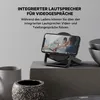 A smartphone is placed on a charging stand, displaying a video call. The scene is set on a table with decorative pots and bowls. Produktbild-Vorschau