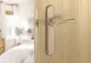 A close-up of a door handle and lock mechanism on a wooden door, with a blurred bedroom interior visible through the open doorway. Produktbild-Vorschau