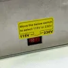A close-up of a voltage selector switch on a metal appliance, with a yellow sticker providing instructions. Podgląd zdjęcia produktu