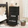 A black fabric dresser with four drawers is placed next to a bed in a bedroom setting. A lamp and a framed photo sit on top of the dresser. A basket of white flowers is on the floor to the right. Podgląd zdjęcia produktu