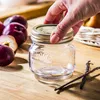 A hand is placing a lid on a small Kilner jar, surrounded by plums and vanilla beans on a wooden surface. Produktbild-Vorschau