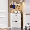 A white shoe cabinet with two drawers and a shelf above, located in an entryway next to a white door. There are hats, keys, and small baskets on the shelf. Anteprima dell’immagine prodotto