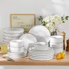 A collection of white ceramic dinnerware with black rim stripes, arranged on a wooden surface with a framed picture and flowers in the background. Voir les images du produit