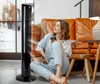 A woman sits on the floor in front of a tower fan, holding a phone. Anteprima dell’immagine prodotto