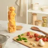 A glass jar filled with pasta sits on a marble countertop next to a cutting board with tomatoes, basil, and garlic. Produktbild-Vorschau