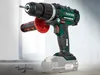 A cordless drill with a battery pack attached, resting on a surface with screws scattered nearby. The background is a blurred interior scene with a window. Produktbild-Vorschau