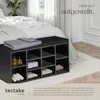 A black storage bench with multiple compartments, placed on a rug in a bedroom next to a bed. The bench is topped with folded blankets. Bottles and folded towels are visible inside some compartments. Náhled obrázku produktu