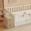 A white wooden children's bed with a mattress and a safety rail. Two beige storage boxes are stacked next to the bed. Náhľad obrázku produktu