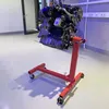 A car engine is mounted on a red engine stand in a workshop or garage setting. Náhľad obrázku produktu