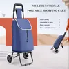 A blue multifunctional portable shopping cart is shown in a studio setting with a decorative orange backdrop and a sandy desert-like background. A person is shown in the background using a similar cart. Produktbild-Vorschau