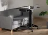 A black adjustable laptop table is positioned next to a gray sofa in a living room setting. The table has a laptop, a glass of water, and a book on its surface. The sofa has wooden legs and is upholstered in gray fabric. The floor is made of light brown wo Produktbild-Vorschau