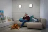 A man is sitting on a couch with headphones on, looking at a tablet, with a dog lying on the floor in front of him. The room has a concrete wall and modern decor. Produktbild-Vorschau