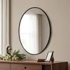A round mirror with a black frame hangs on a wall above a wooden dresser. There are decorative items on the dresser, including flowers and a picture frame. Produktbild-Vorschau