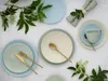 A table setting with pastel-colored plates, bowls, and a cup, accented by greenery and golden cutlery. Náhľad obrázku produktu