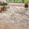 A rug with a marble-like pattern is laid out on a wooden floor in a living room setting. A gray armchair with cushions and a yellow throw is placed on the left side of the rug. A window with a view of green trees is visible in the background. Voir les images du produit