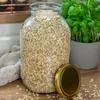 A large glass jar filled with oats sits on a wooden surface next to a gold lid and a potted plant. Podgląd zdjęcia produktu