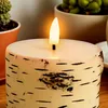 A close-up of an LED candle with a birch bark texture, placed on a wooden surface with potted plants in the background. Produktbild-Vorschau