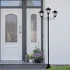 A black solar-powered lamppost with three lights stands on a porch next to a white brick wall and a double white door. Produktbild-Vorschau