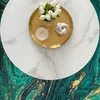 A close-up view of a round marble-patterned table with decorative items on it, placed on a green and gold abstract patterned rug. Náhled obrázku produktu