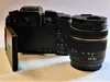 A black digital camera with a lens attached, shown from the back with the screen open. Produktbild-Vorschau
