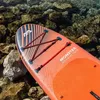 The nose of an orange stand-up paddleboard resting on a rocky shore with clear water in the background. Produktbild-Vorschau