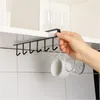 A hand is holding a black under-shelf mug rack with hooks, which is attached to the underside of a white cabinet. Two mugs and a glass are hanging from the hooks. Produktbild-Vorschau
