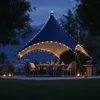 An outdoor dining area at night, set up under a large tent with string lights, featuring a table set for dinner and a grill. Produktbild-Vorschau
