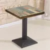 A square table with a multi-colored wood-grain top and a dark metal pedestal base, set against a textured wall and a white floor. Produktbild-Vorschau