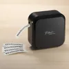A black label printer is shown on a wooden surface, printing out several strips of paper with the word "Welcome!". Náhled obrázku produktu