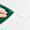 A hand is wiping water droplets off a white surface with a green microfiber cloth. Produktbild-Vorschau