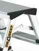 A close-up view of the top section of a silver aluminum step ladder, showing a black plastic protective piece and a yellow sticker with black text. Produktbild-Vorschau