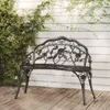 A black metal garden bench with rose detailing sits on a wooden deck in front of a white paneled wall with ivy growing on it. Náhled obrázku produktu