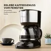 A coffee maker is shown on a countertop with a cup and saucer, and a small plant in the background. Produktbild-Vorschau