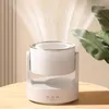 A white humidifier with a gold rim is shown on a wooden table, emitting a mist. A large green leaf is visible to the right. Produktbild-Vorschau