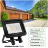 A black LED floodlight is shown in the foreground, with a brick house with three garage doors in the background. Voir les images du produit