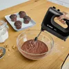 A hand is using a black Bosch hand mixer to mix chocolate batter in a glass bowl on a wooden table. There are already baked chocolate muffins on a tray in the background, and a jar of flour and walnuts on the table. Produktbild-Vorschau