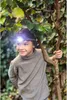 A young boy wearing a headlamp stands in a natural outdoor setting, leaning against a tree branch. Produktbild-Vorschau