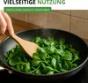A hand is using a wooden spatula to stir fresh green leaves in a black frying pan on a stovetop. Produktbild-Vorschau