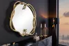 A decorative snake-shaped mirror is displayed on a dark wall above a black dresser. Two candlesticks with lit candles and a small perfume bottle are also on the dresser. A floor lamp is visible to the right, and a cityscape can be seen through a window in  Produktbild-Vorschau