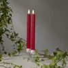 Two red LED taper candles with a flame-like top are placed in clear glass candle holders, surrounded by green ivy on a white surface against a gray background. Produktbild-Vorschau