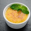 A close-up of a crème brûlée in a white ramekin, garnished with a mint leaf, resting on a dark textured surface. Anteprima dell’immagine prodotto
