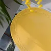 A close-up view of a yellow oval plate with a decorative white dashed line pattern around its rim, placed on a surface with some greenery and yellow flowers. Náhľad obrázku produktu