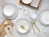 A flat lay of white ceramic dinnerware with brown trim, including plates, bowls, and a mug, arranged on a light gray surface with some decorative elements like dried grass and a wooden board. Náhľad obrázku produktu