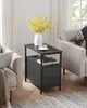 A dark gray side table with two drawers is placed next to a light gray armchair in a living room setting. A white vase with yellow flowers, a cup of coffee, and a remote are on top of the table. Someone is sitting in the armchair on the left, using a lapto Náhled obrázku produktu