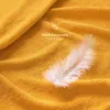 A close-up of a yellow textured fabric, likely a bedsheet, with a single white feather resting on it. Text overlays indicate softness and coziness. Anteprima dell’immagine prodotto